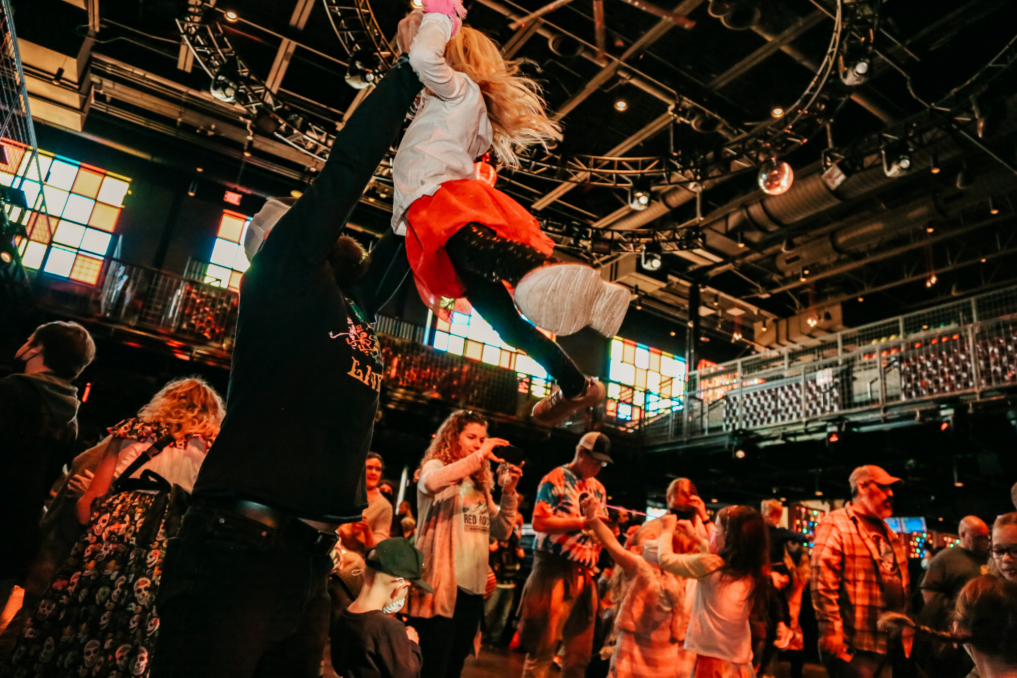 A crowded indoor venue with people dancing; an adult lifts a child in the air, and colorful lights fill the space.