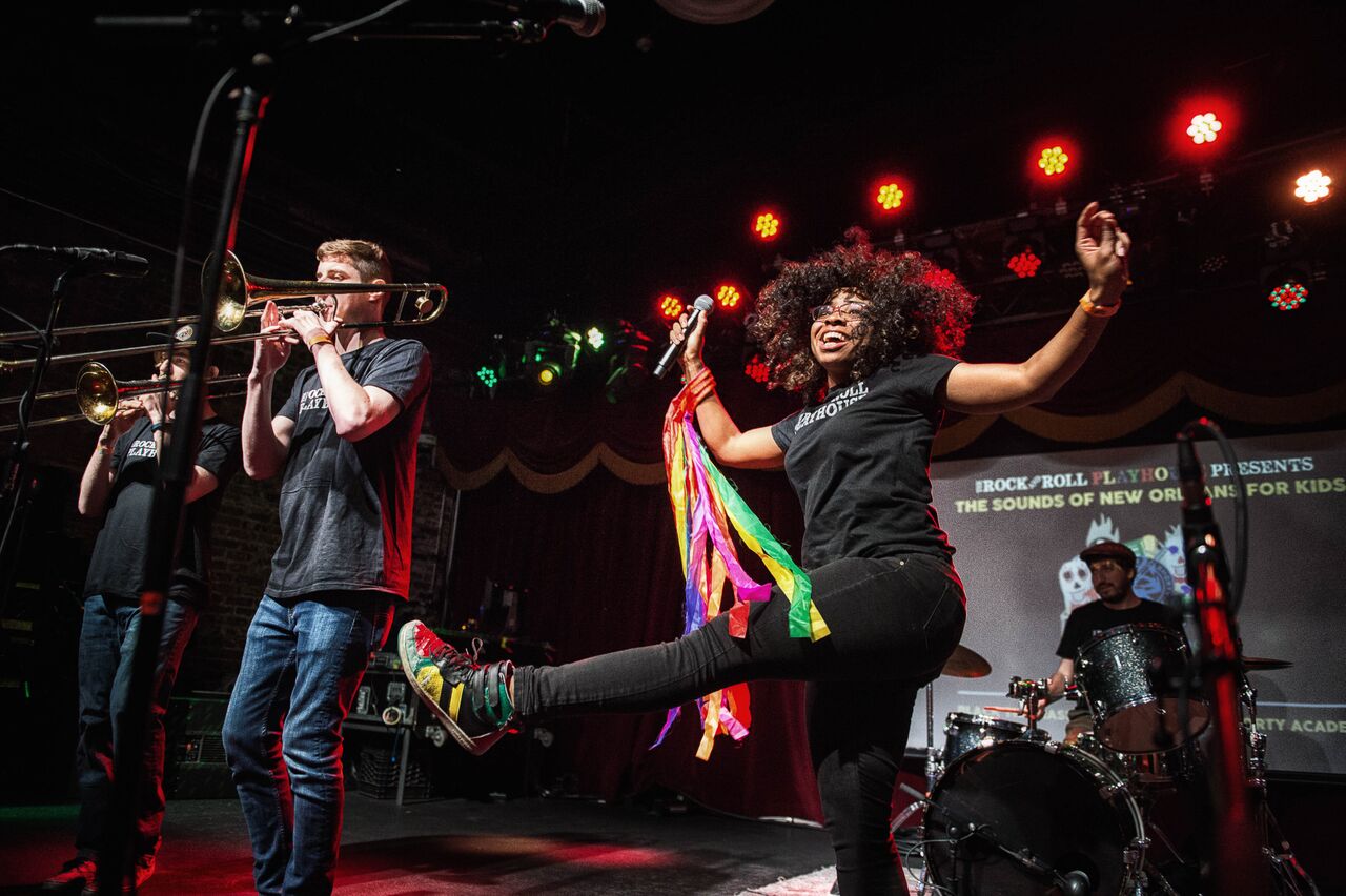A band performs on stage; a woman sings and dances with colorful ribbons while a trombone player and drummer accompany her under stage lights.