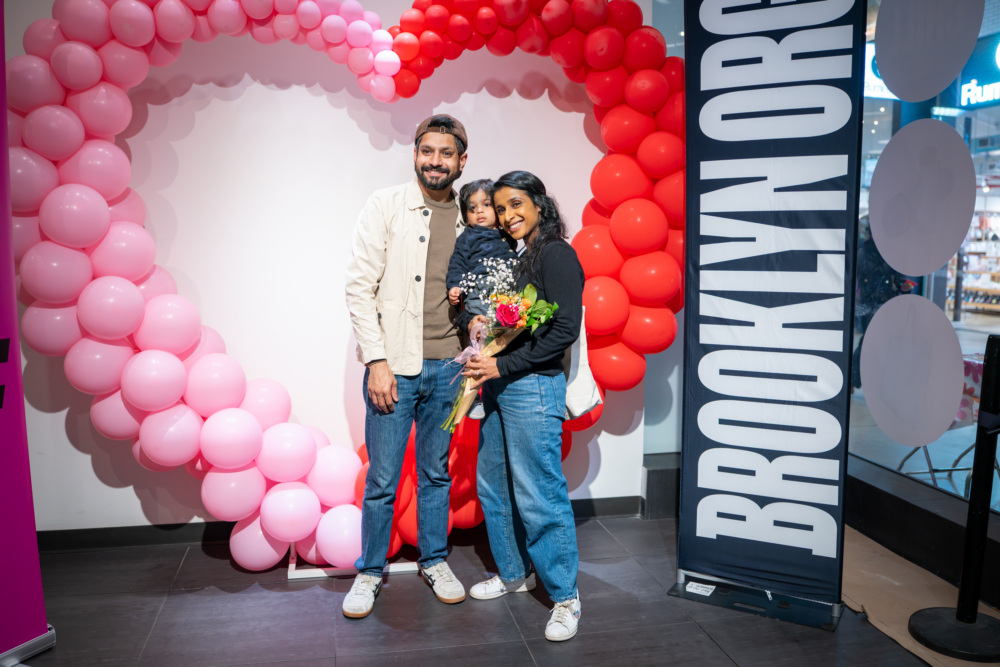 A family of three stands in front of a pink and red balloon heart display and a "Brooklyn Org" banner; the woman holds flowers and the child.