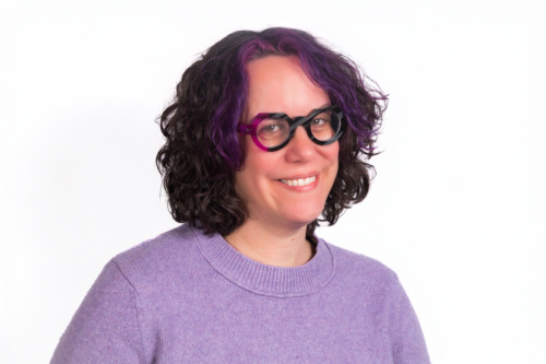 A person with curly dark hair and purple streaks, wearing round black and purple glasses and a light purple sweater, smiles at the camera against a white background.