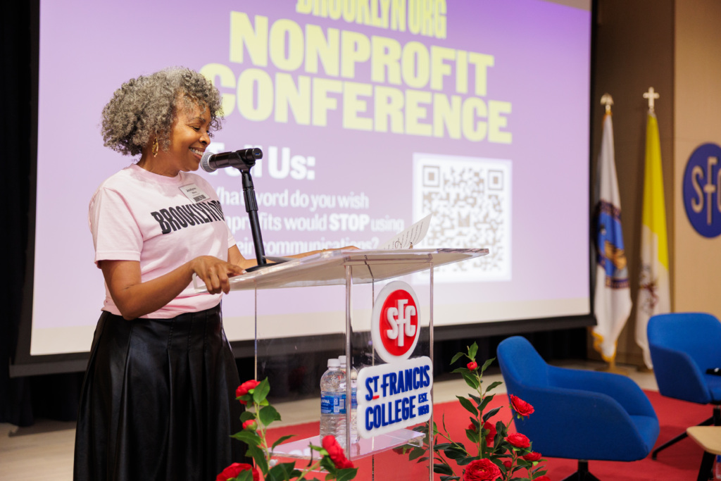 A woman speaks at a podium during the Brooklyn.Org Nonprofit Conference at St. Francis College; a projector screen and chairs are visible in the background.