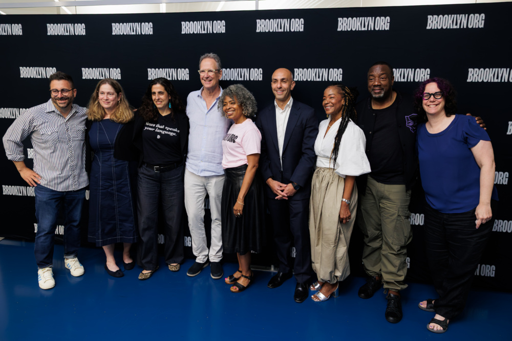 Nine people stand side by side, smiling for a group photo in front of a step-and-repeat backdrop with “BROOKLYN.ORG” printed on it.
