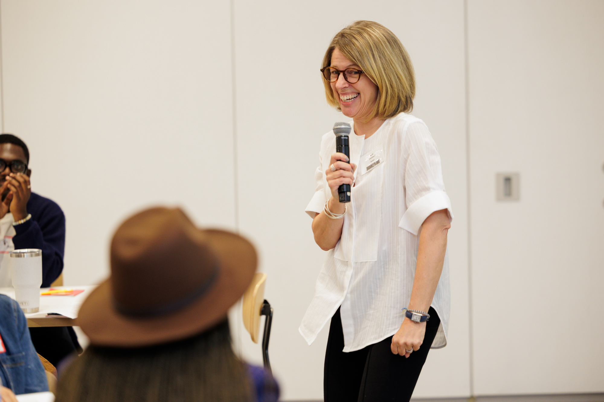 A woman in glasses holds a microphone and smiles while speaking to a seated group in a brightly lit room.