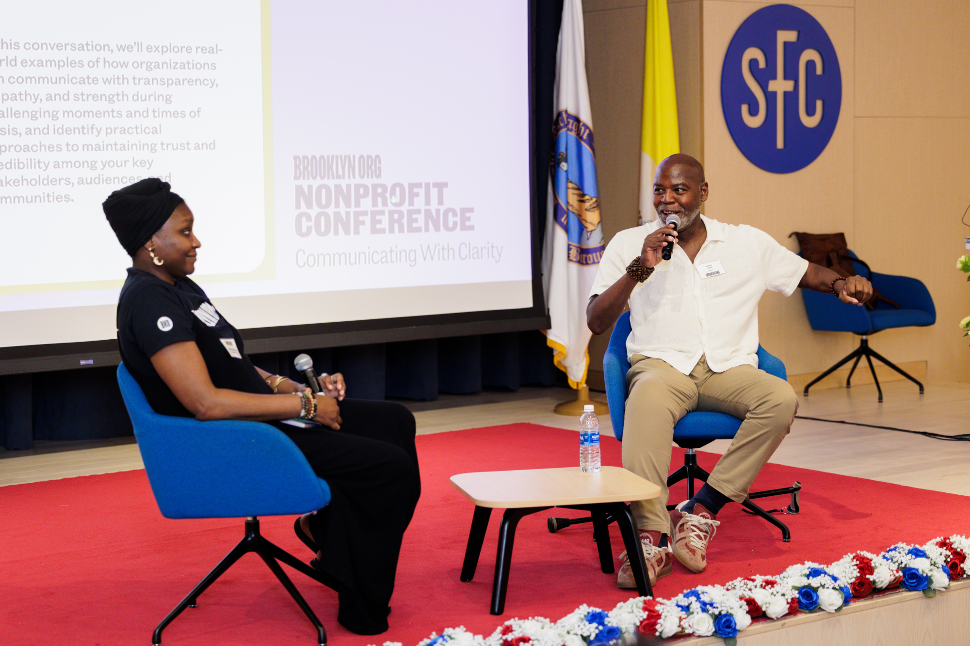 Two people sit on stage with microphones during a nonprofit conference, with a presentation screen and organization banners in the background.