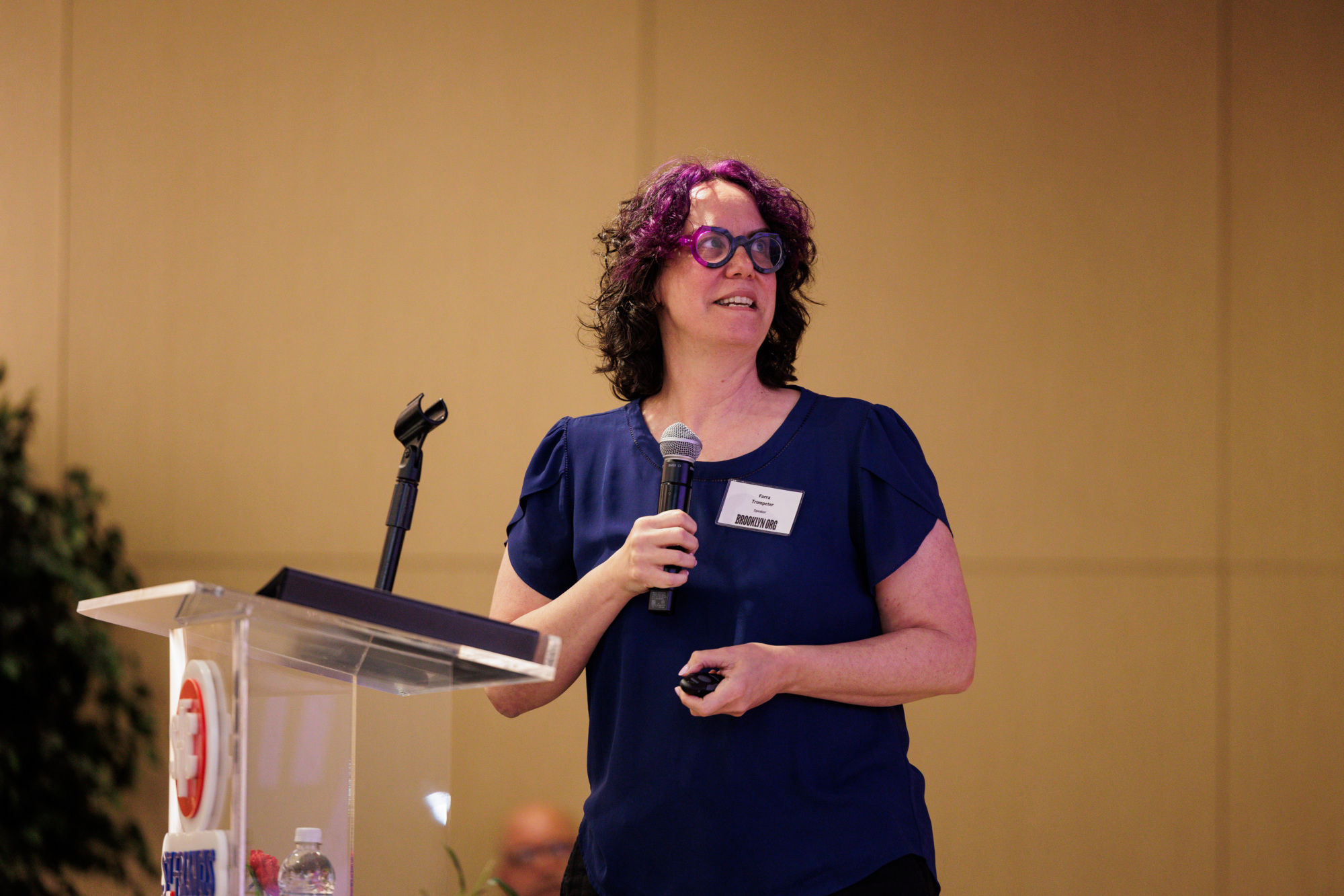 A person with curly, purple hair and glasses stands at a podium, holding a microphone and a remote, giving a presentation.