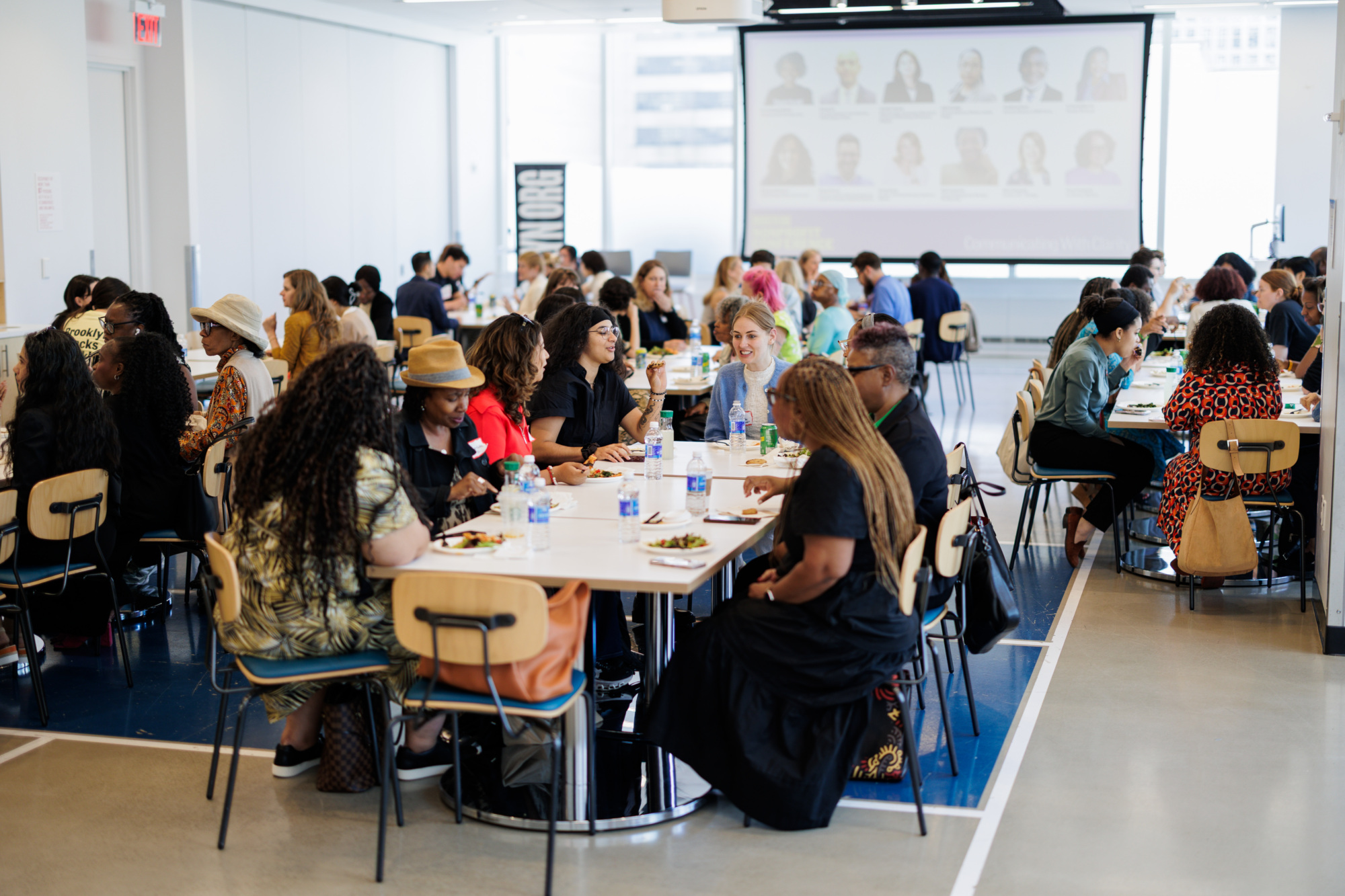 A group of people sit at tables eating and talking in a large, bright conference room with a presentation screen at the front.