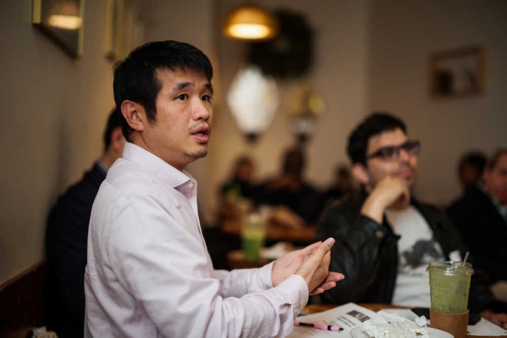A man in a light shirt gestures while speaking during a meeting, with other people seated and listening in the background. Papers and a drink are on the table in front of him.