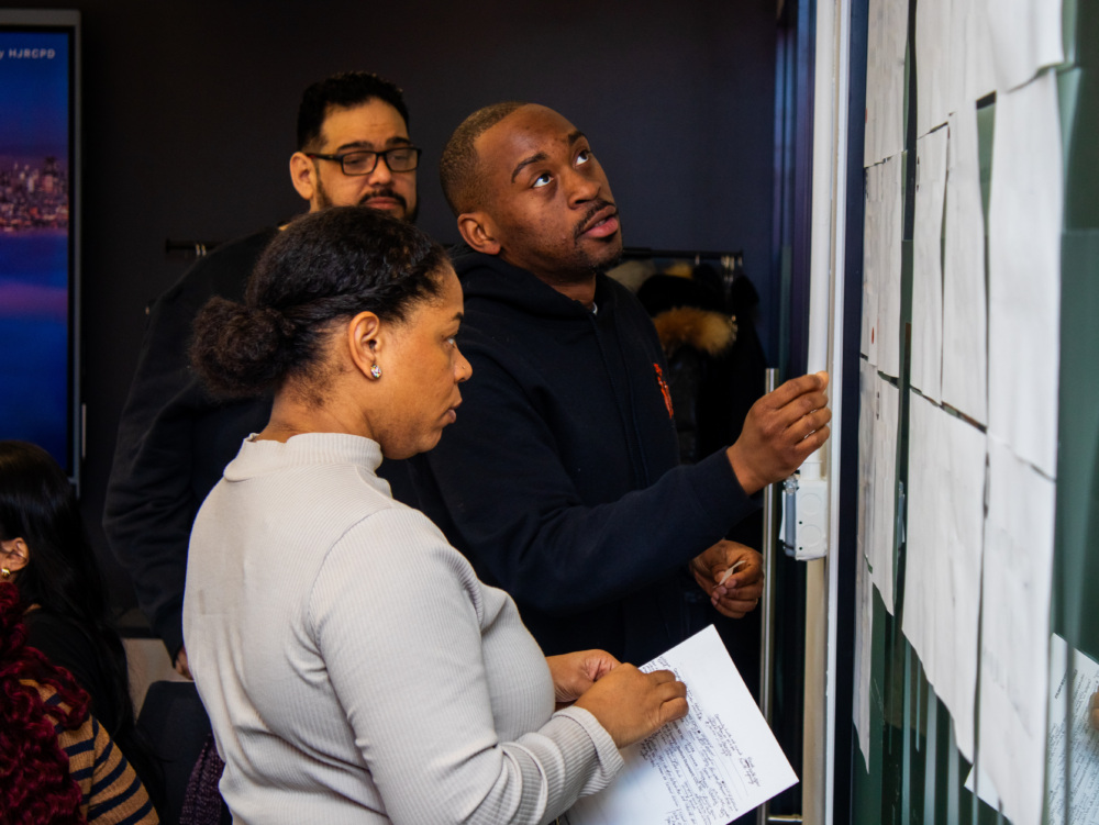 Three people stand in front of a wall covered with papers, discussing and examining the documents; one person holds a sheet while another points at the wall.