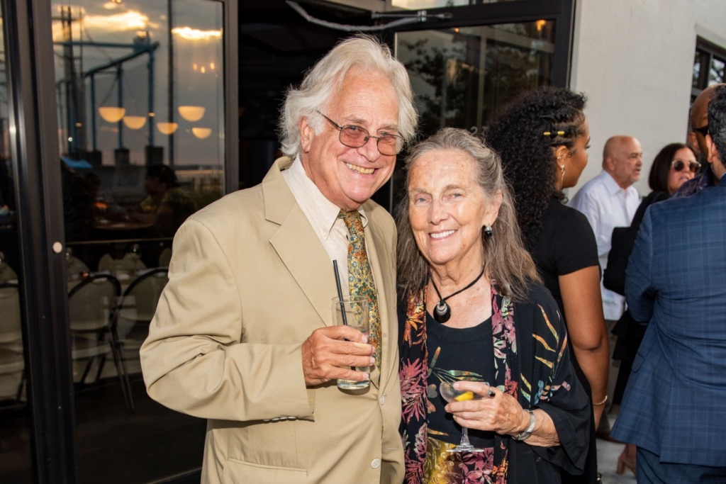 An older man in a tan suit and an older woman in a black floral dress stand together outside, smiling and holding drinks at a social event.