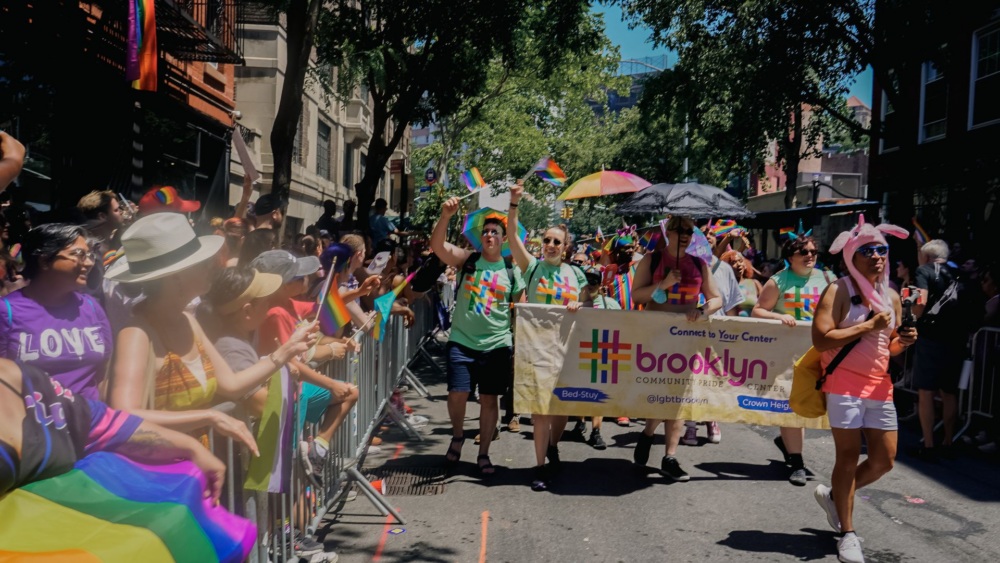 A group of people march in a pride parade holding a "Brooklyn Community Pride Center" banner, surrounded by cheering spectators with rainbow flags.