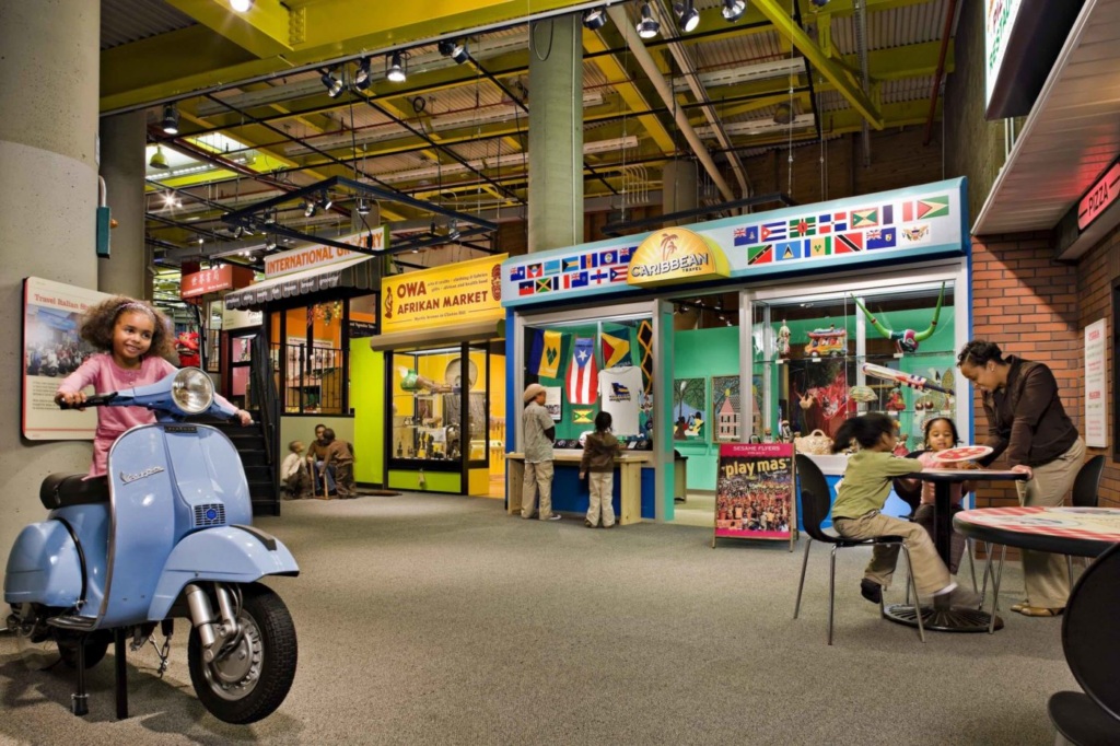 Children and adults engage in various activities in a colorful, multicultural exhibit area with flags, displays, and a blue scooter indoors.