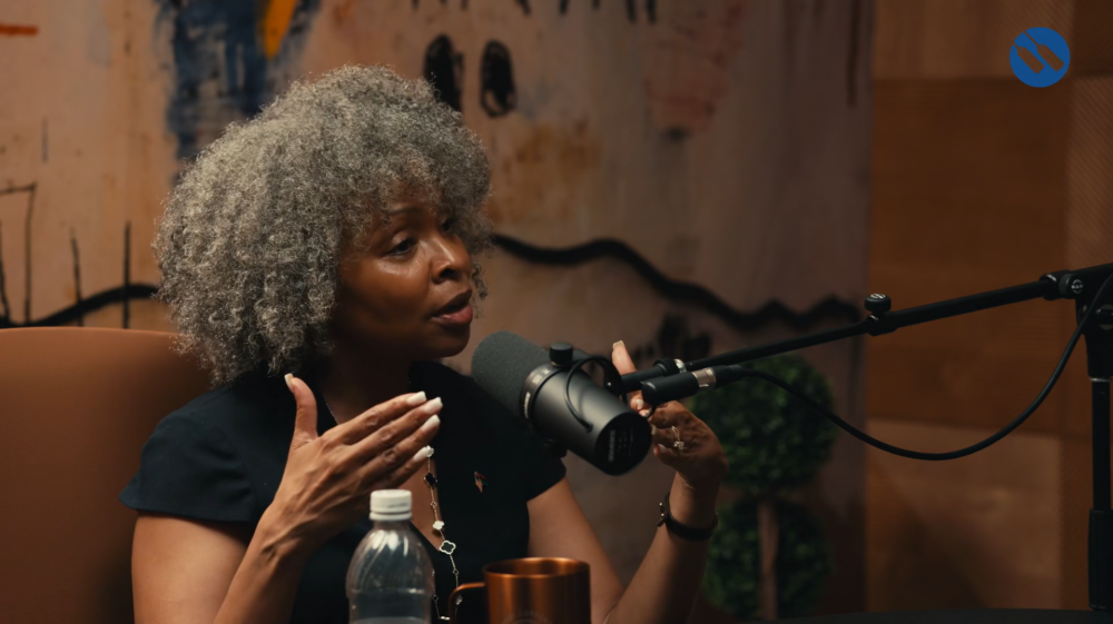 A woman with gray curly hair speaks into a microphone during a podcast or interview, gesturing with her hands. A water bottle and cup are on the table in front of her.