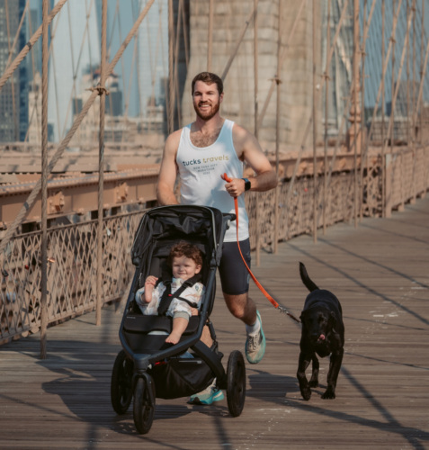 A man jogs across a bridge while pushing a stroller with a child and holding the leash of a black dog. City buildings are visible in the background.