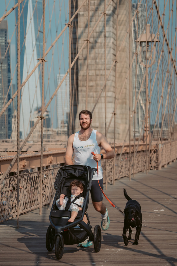 A man jogs on a bridge while pushing a stroller with a child inside and walking a black dog on a leash.
