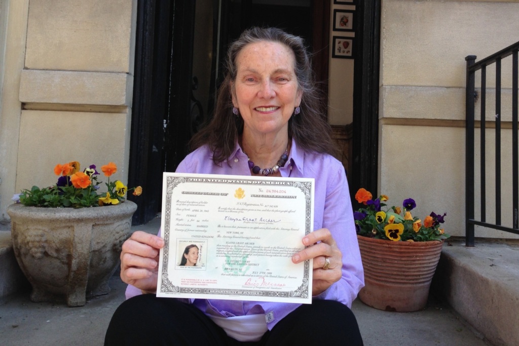 Older woman sitting on steps outdoors, smiling and holding up an official certificate with a photo and text. Potted flowers are visible on either side of her.