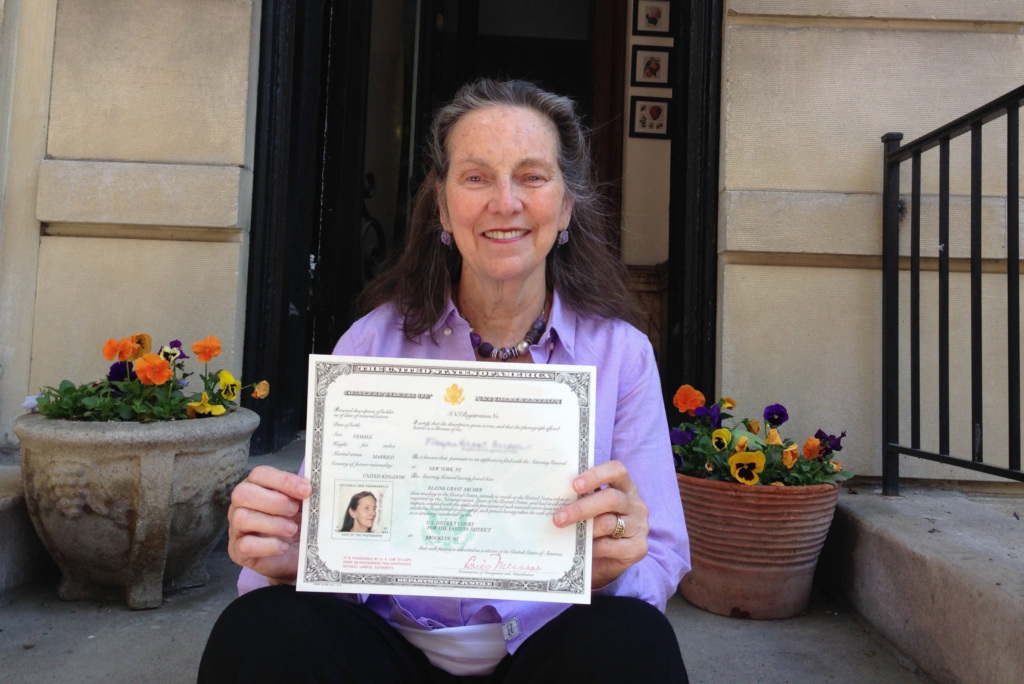 Older woman sitting on steps outdoors, smiling and holding up an official certificate with a photo and text. Potted flowers are visible on either side of her.