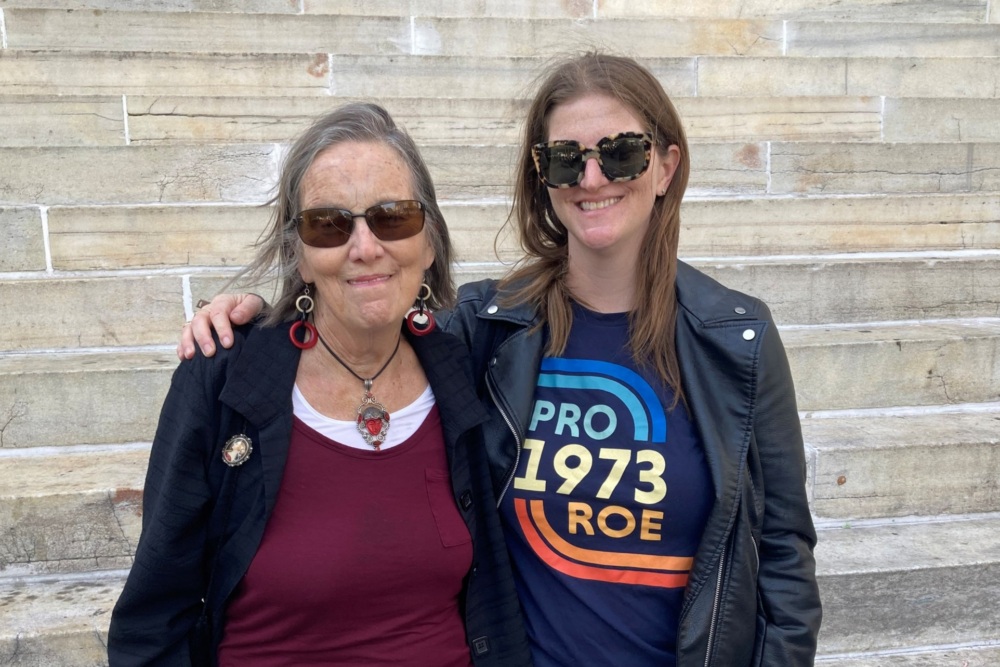 Two people with sunglasses pose on stone steps. One wears a "Pro Roe 1973" t-shirt and a leather jacket, while the other wears a maroon shirt and black jacket.