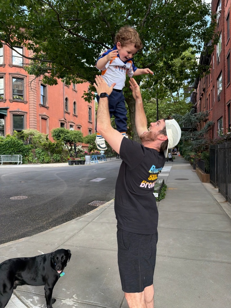 A man lifts a smiling toddler into the air on a city sidewalk, with a black dog standing nearby and brick buildings in the background.