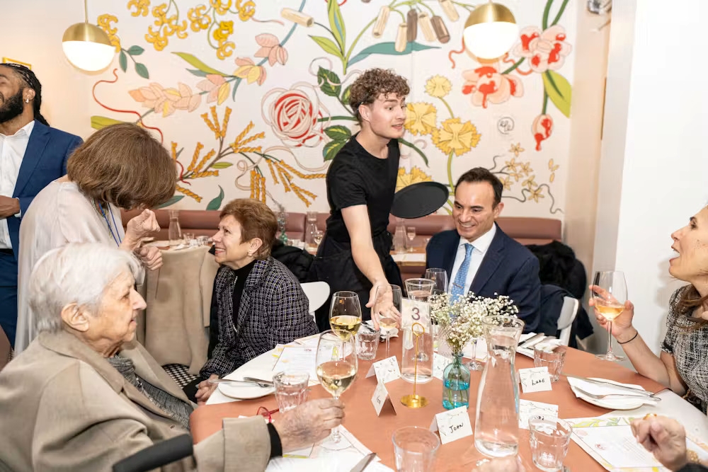 A group of people sits and stands around a restaurant table with drinks, menus, and name cards, engaging in conversation under a floral mural.
