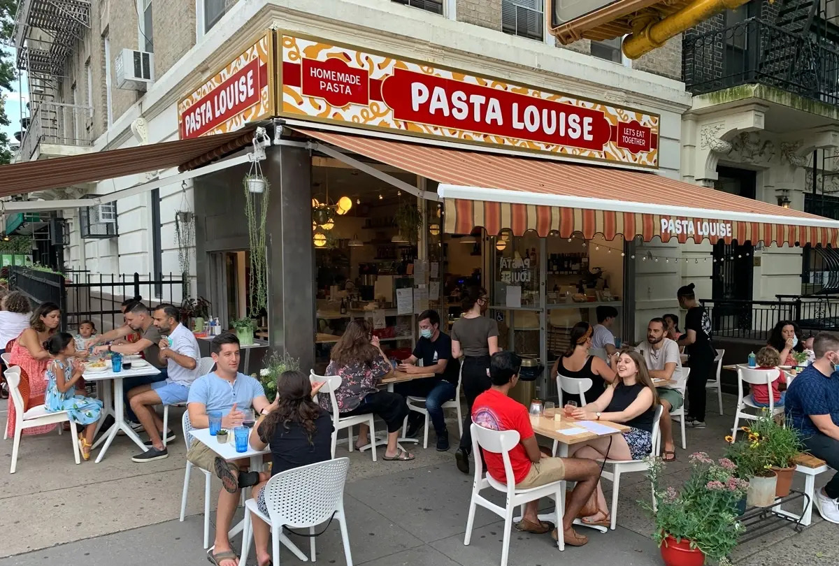 People are dining outdoors at Pasta Louise, a restaurant with a striped awning and signage advertising homemade pasta, on a city sidewalk.