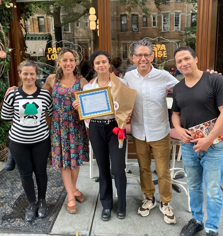 Five people stand outside a restaurant; one person in the center holds a certificate and a bouquet of flowers, while the others smile at the camera.