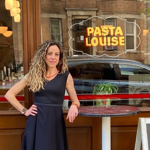 A woman in a black dress stands beside a small table outside a restaurant with a "PASTA LOUISE" sign on the window.