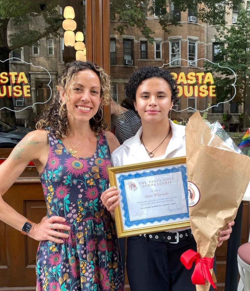 Two people stand outside a restaurant. One holds a bouquet and a framed certificate. They smile at the camera. The restaurant window displays the words "PASTA LOUISE.