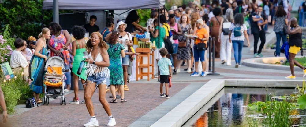 A crowd of people gathers outdoors near a pond and pathway, with some standing in line, others walking, and a few pushing strollers.