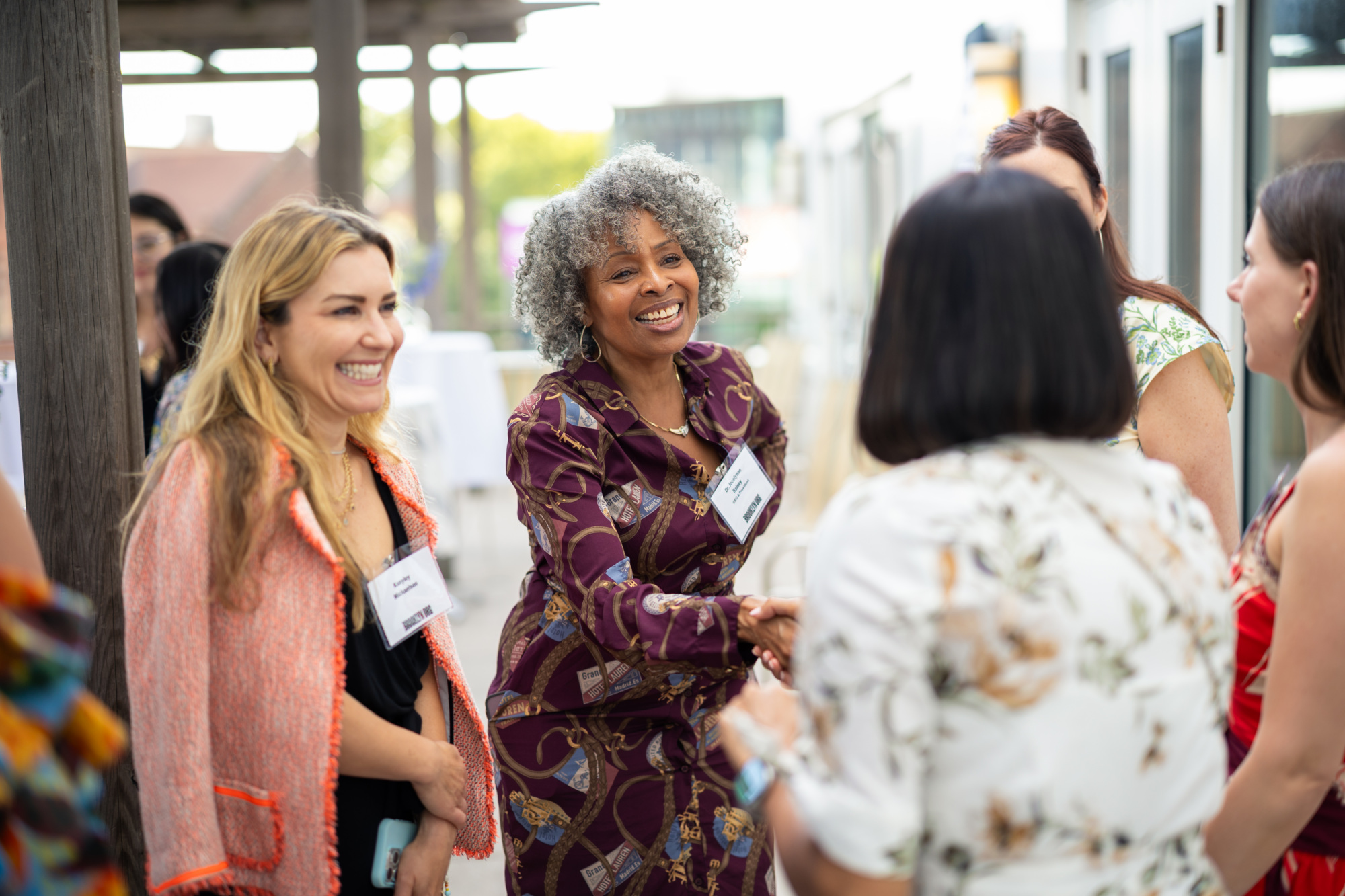 A group of women, some wearing name tags, stand and interact at an outdoor event; one woman in patterned attire is shaking hands and smiling.
