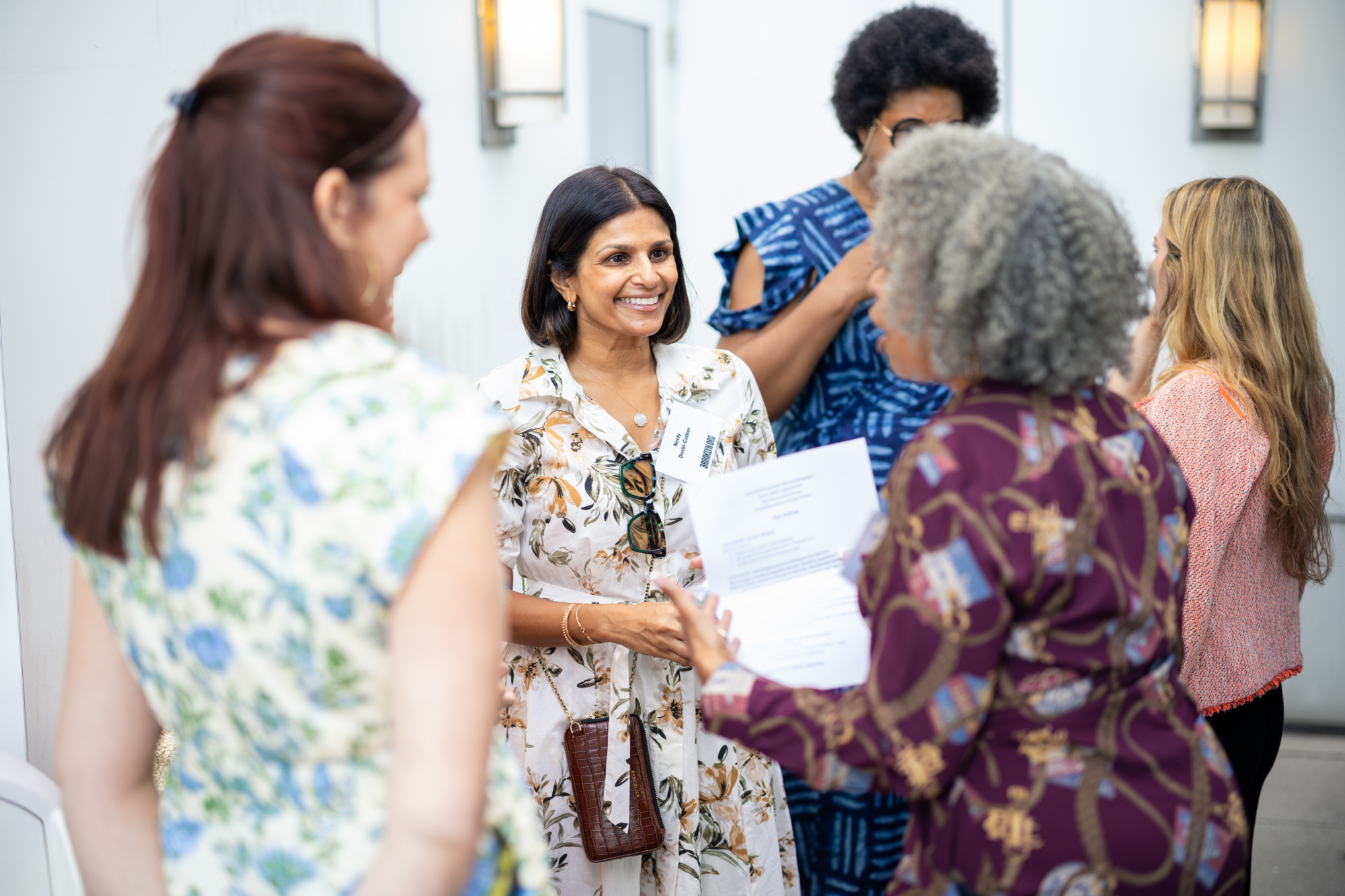 A group of women engage in conversation indoors, with one woman holding a document and others smiling and listening.