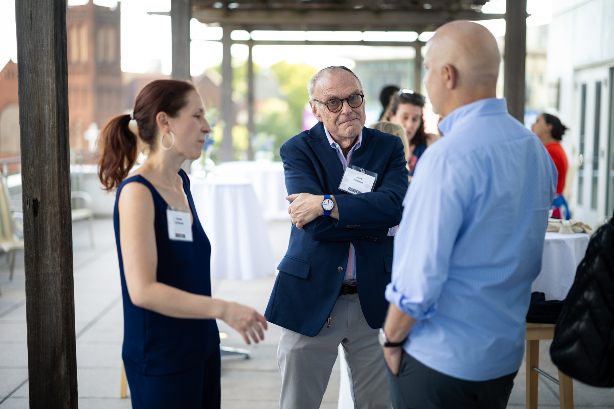 Three adults stand on a patio having a conversation during a daytime event; other people and tables are visible in the background.