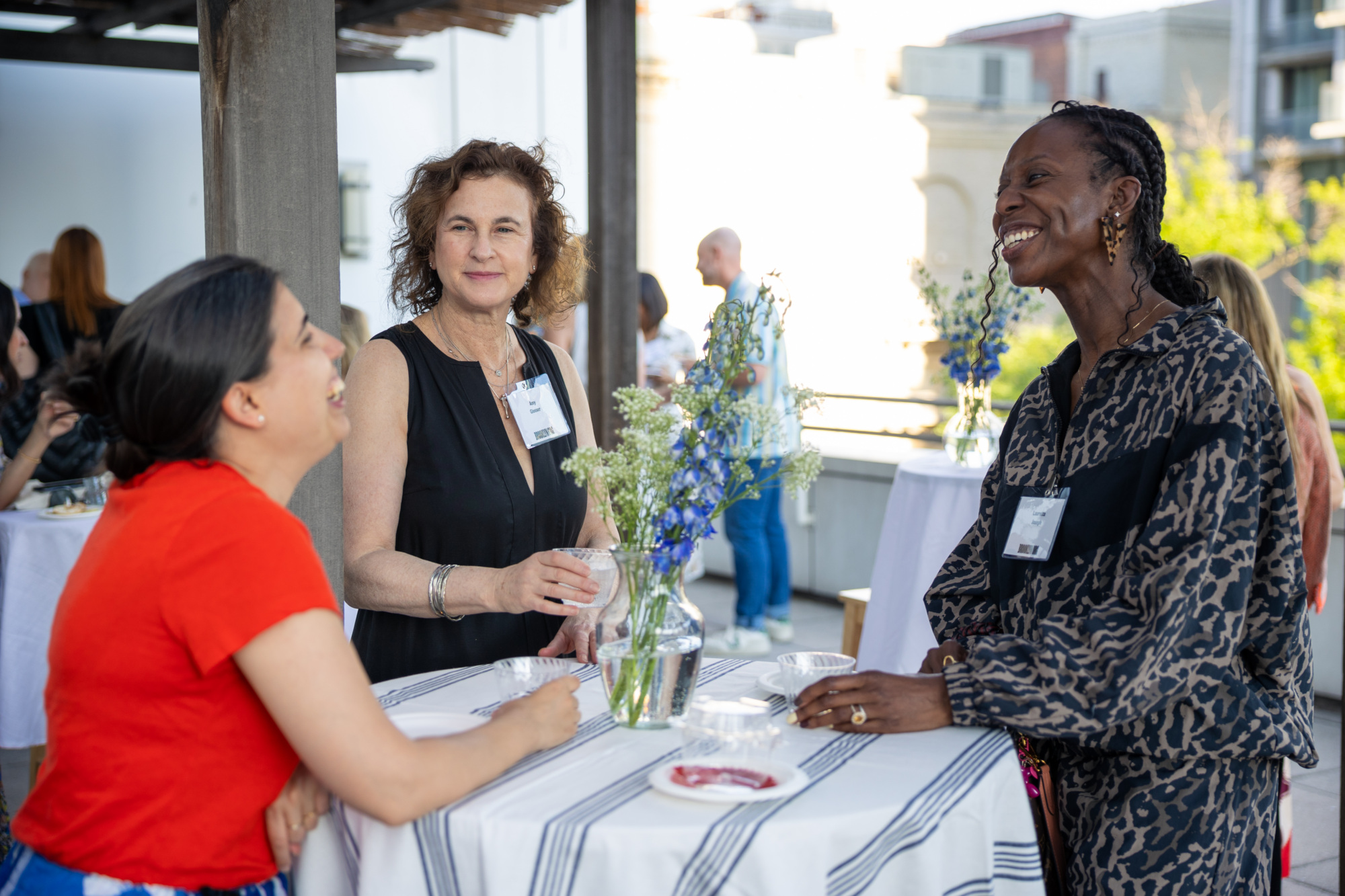 Three women stand and talk around a small table with flowers at an outdoor event, smiling and engaged in conversation.