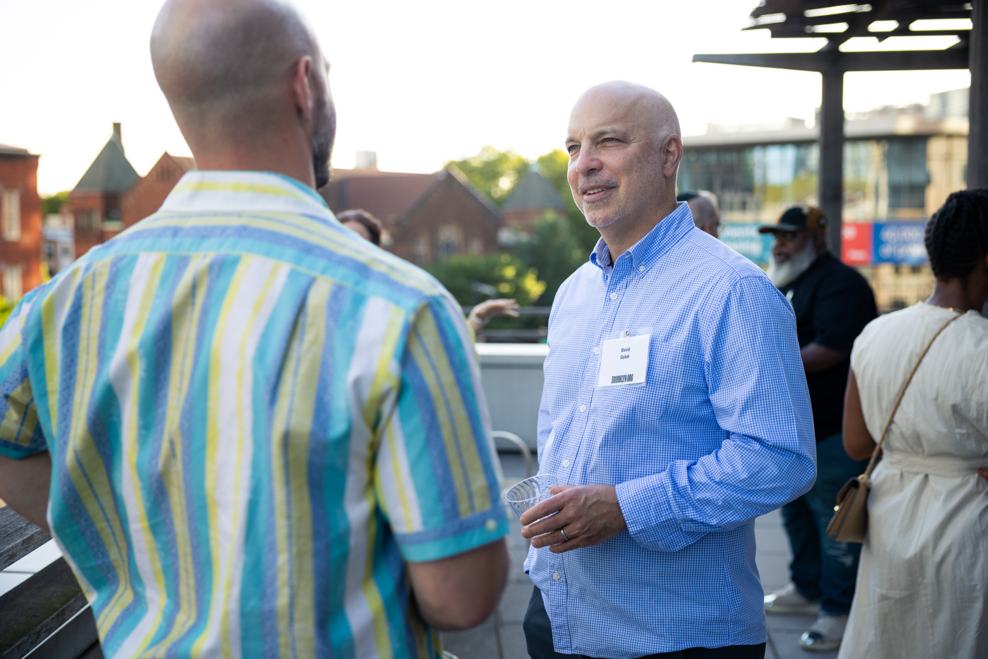 A group of people converse outdoors at a social event; one man in a blue shirt holds a drink and wears a name tag.