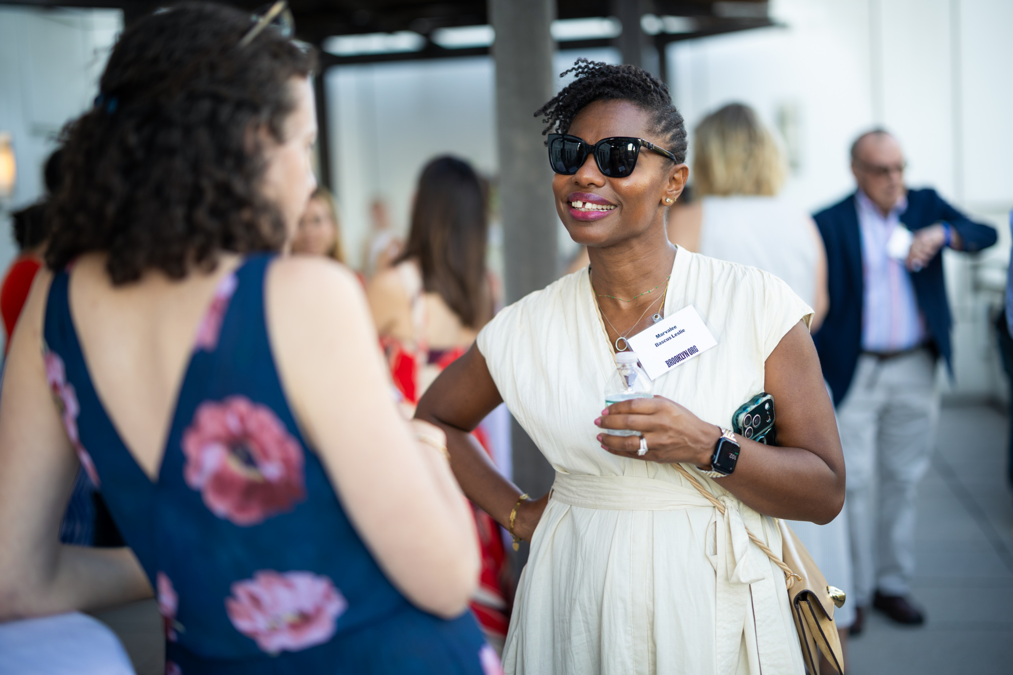 Two women converse at an outdoor event; one wears sunglasses and a white dress, holding a drink and phone. People socialize in the background.