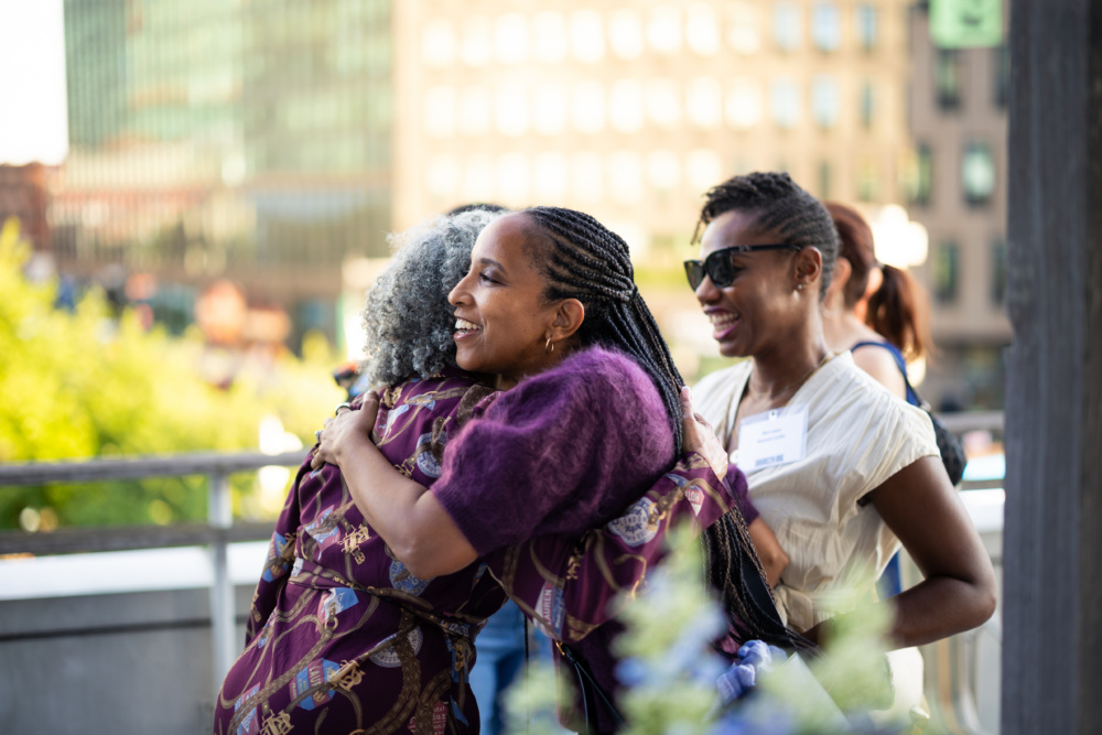 Two women embrace outdoors while another woman smiles nearby. Buildings and greenery are visible in the background on a sunny day.