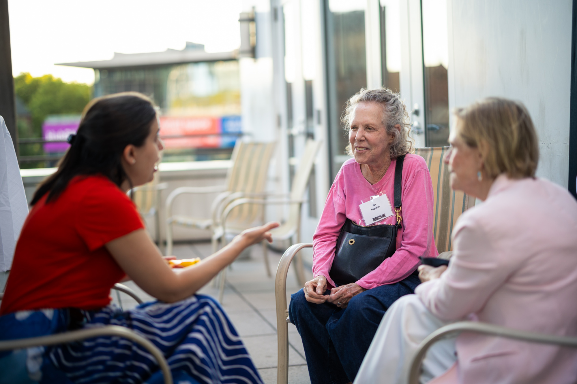 Three women sit outdoors on a patio, engaged in conversation. One gestures while talking, and the other two listen attentively.