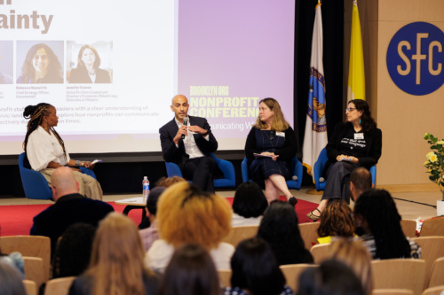 Four panelists sit on stage at a nonprofit conference, engaging in a lively discussion about nonprofit training while speaking to an attentive audience. Behind them is a projected slide with names, photos, and event branding.