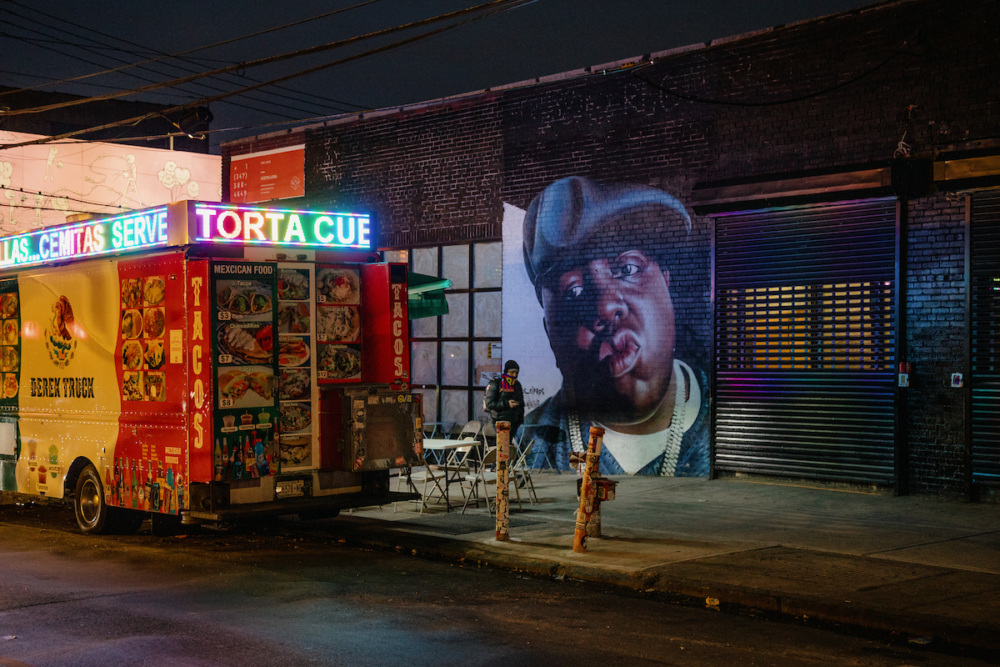 A brightly lit food truck is parked on a dim street at night beside a large mural of a man in a hat on a brick wall, with a person standing nearby.