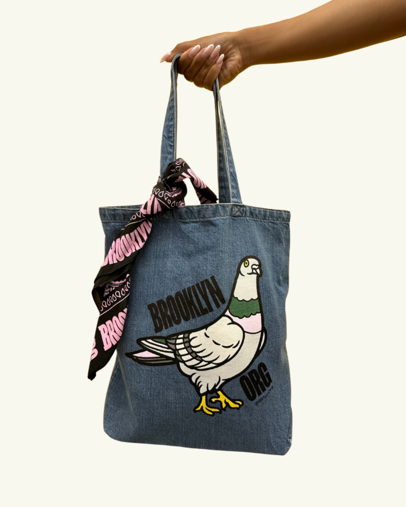 A hand holds a denim tote bag featuring a pigeon graphic, the word “Brooklyn,” and a pink and black scarf tied to one handle.
