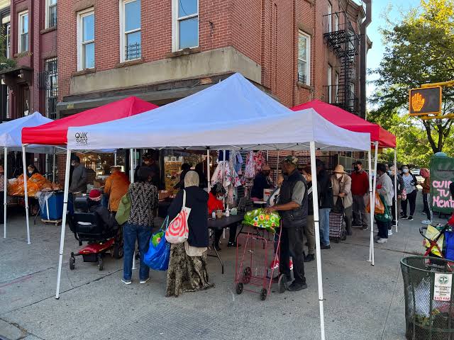 People gather under canopies at a sidewalk market on a city street corner. Various items, groceries, and clothing are displayed for sale. Brick buildings and a pedestrian signal are visible.