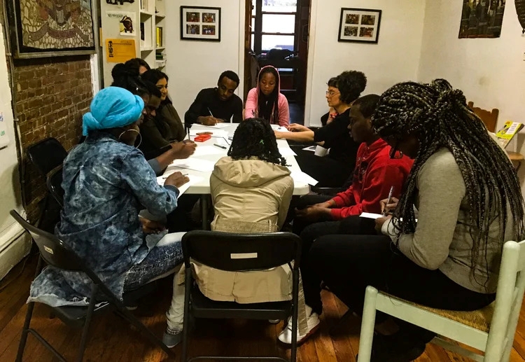 A group of people sit around a table in a room, engaged in discussion and taking notes, with bookshelves and framed photos on the walls.