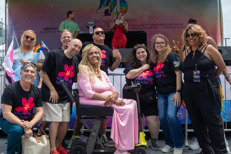 A group of people pose and smile at an outdoor event, with one person in a wheelchair in the center, and rainbow decorations and a stage in the background.