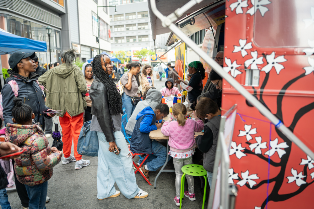 People gather around a food truck with cherry blossom designs while children sit at a table engaging in activities at a busy outdoor event.