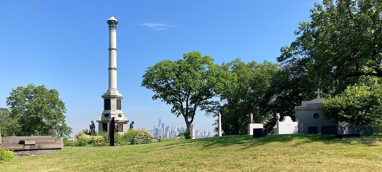 A grassy cemetery with a tall column monument, trees, and gravestones; a city skyline is visible in the distant background under a clear blue sky.