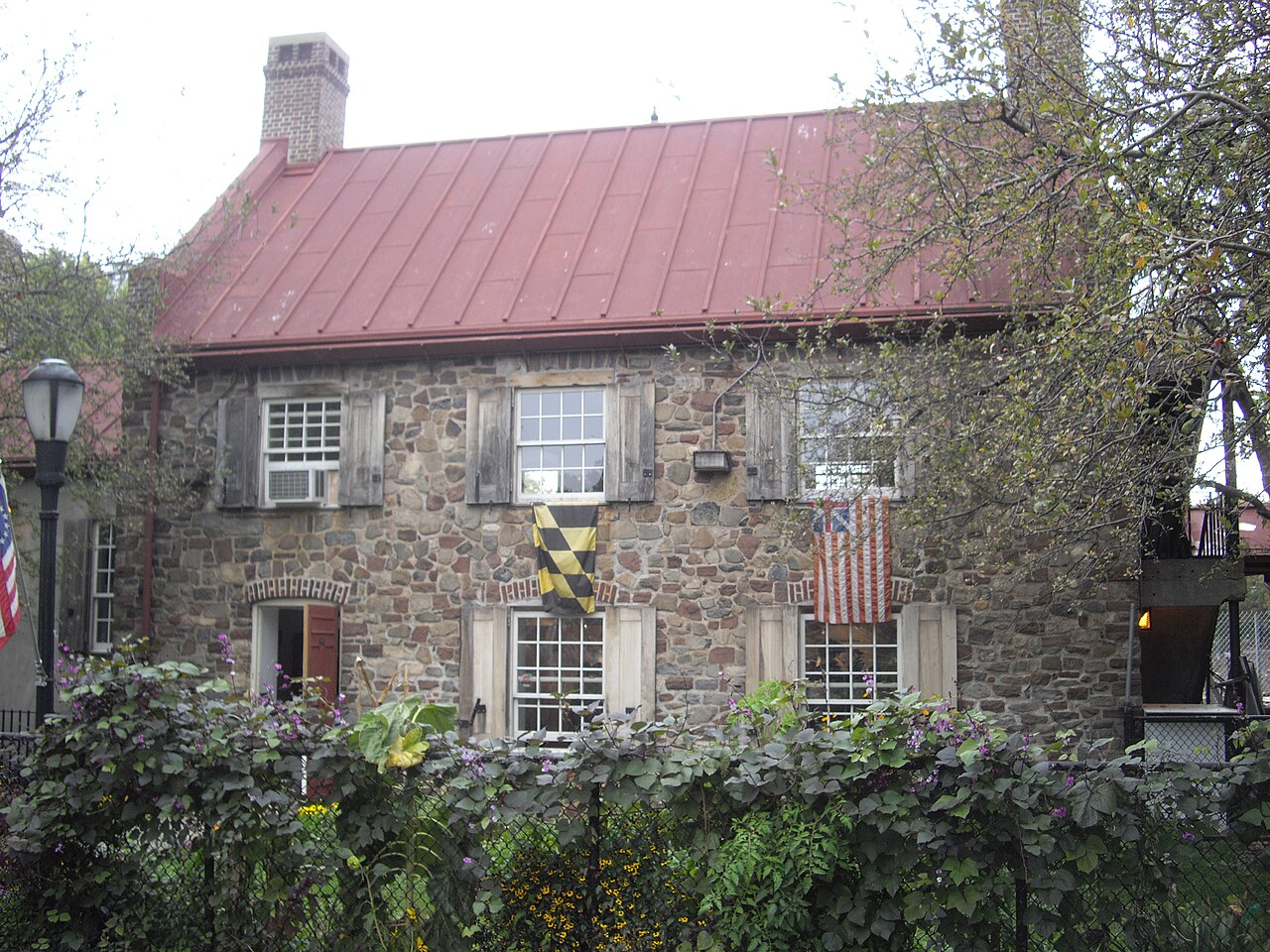 Stone house with a red roof, two flags (Maryland and U.S.) hanging from upstairs windows, partly obscured by a green leafy fence and trees.