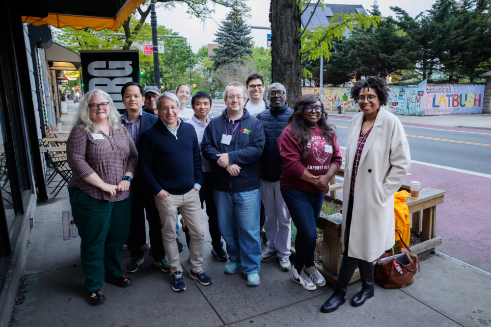 A group of eleven adults stands together on a city sidewalk, posing for a photo near outdoor tables and a mural reading “LATBUSH” in the background.