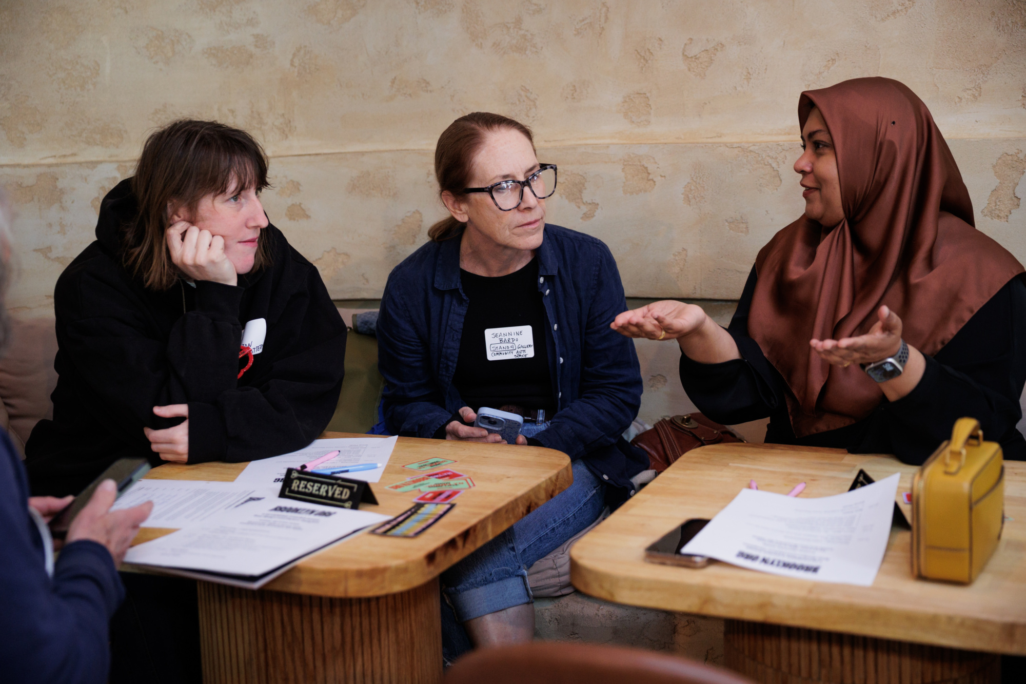 Three women sit at a table in conversation with papers, pens, and a “Reserved” sign in front of them in a casual indoor setting.