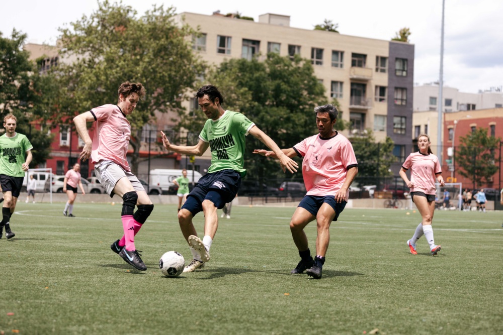 Three soccer players compete for the ball on a grass field, with other players and apartment buildings visible in the background.