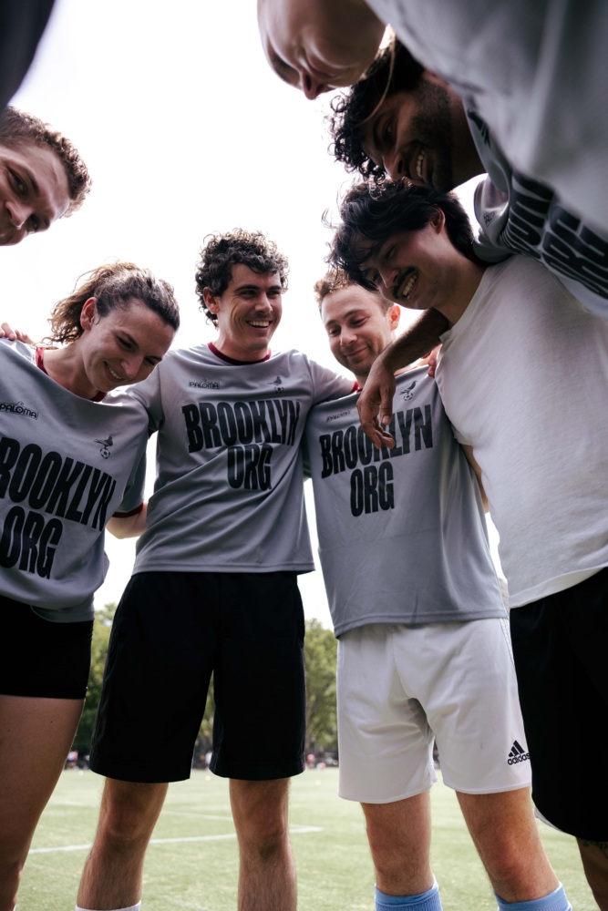 A group of six soccer players in Brooklyn Org shirts stand in a huddle on a field, smiling and preparing for a game.