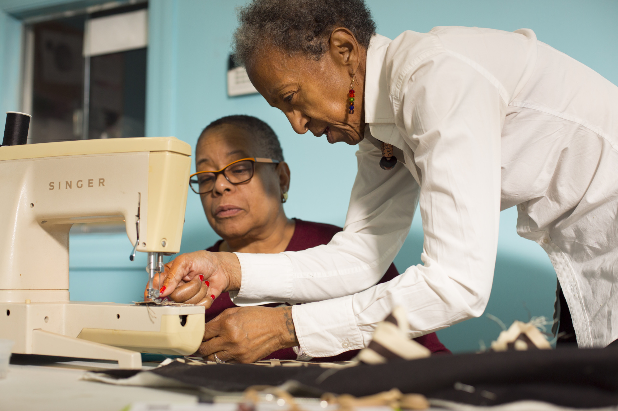 Two women work together at a sewing machine, with one guiding fabric and the other observing closely in a brightly lit room.
